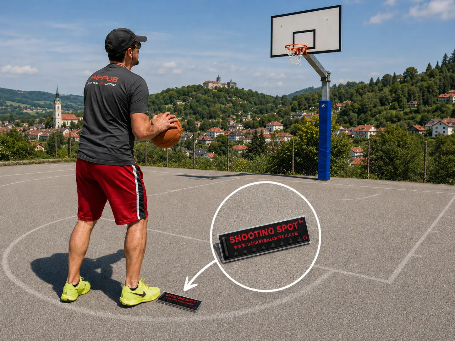 Outdoor basketball scene showing the Basketball Witch black and red Shooting Spot Marker placed on a court near a player, with a hoop and scenic Novo Mesto hills in the background.