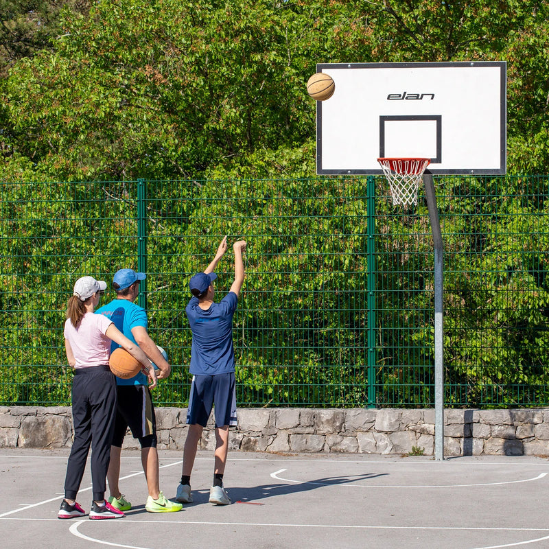 Three people playing basketball on an outdoor court with a green fence and trees in the background.