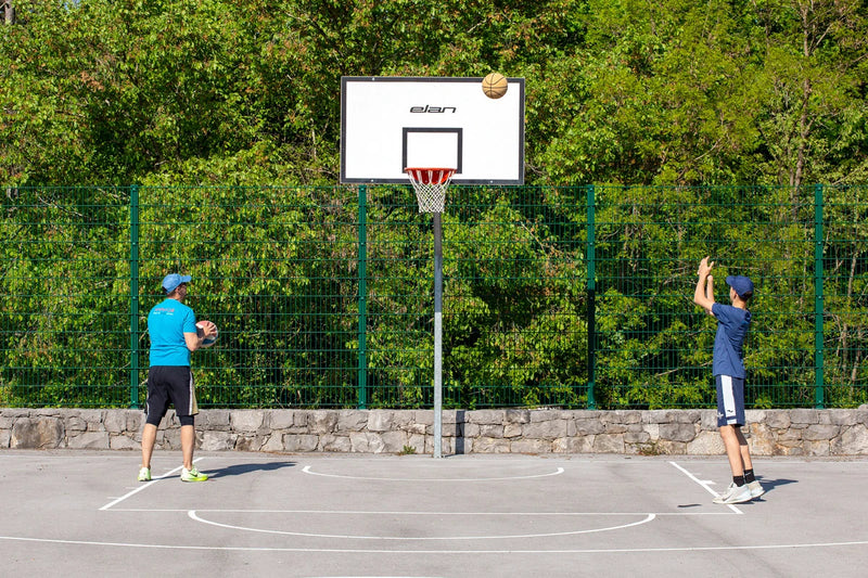 Two people playing basketball on an outdoor court with a green fence and trees in the background.